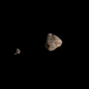 Dinkinesh, a diamond-shaped asteroid, is slightly off center, with a smaller asteroid, Selam, on the left side of the image. Both asteroids are false color edited to be a tan/beige color. They are lit from the right side