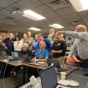 Many people stand around tables spanning across the room, while looking off to the left and pointing to what they see. Many people have shocked faces. Not pictured: the first image of Dinkinesh's not previously known satellite, Selam.
