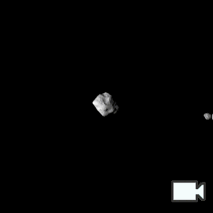 Dinkinesh, a diamond-shaped asteroid in the center, and Selam, a smaller asteroid of two connected rocks on the left, approach, pass across, then depart away from the camera. As they approach, the shape of both asteroids get clearer and the lighting is harsh from the right. As the camera passes across them, Selam moves behind Dinkinesh to the right of it. As they depart, the binary shape of Selam (two connected rocks separated by a black line) is clear, and the lighting comes from the left.