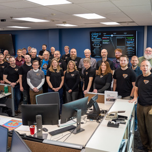 Many smiling people stand in three rows that span across the center of the image, the bottom third being a desk with computer monitors, and the top third being tv screens and the ceiling. Many people are wearing shirts with the Lucy emblem on them.