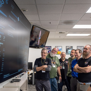 Two tv screens take up 1/3 of the image on the left hand side, on them are updates on the Lucy mission, while many people stand looking and smiling up at the screens.