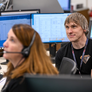 Two people are looking to the left and sitting in from of computer monitors. The person on the left side has a headset on, and the person on the right is wearing a Lucy emblem shirt and lanyard.