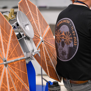 A model of the Lucy spacecraft takes up the left half of the image, the right half is the back of a person wearing a black t-shirt with the Dinkinesh emblem badge.